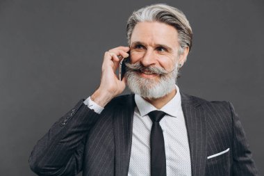 İş ve modaya uygun sakallı, gri takım elbiseli olgun bir adam telefonla konuşuyor ve gri arka planda gülümsüyor..