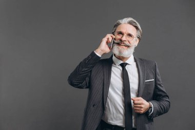 İş ve modaya uygun sakallı, gri takım elbiseli olgun bir adam gri arka planda telefonla gülümsüyor ve konuşuyor..