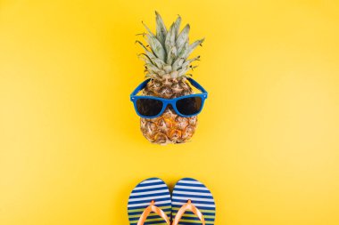 Yaz kavramı. Güneş gözlüklü sevimli ve komik ananas ve sarı arka planda parmak arası terlik.. 