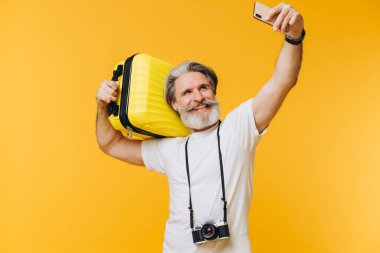 Şık sakallı ve bıyıklı bir adam. Gülümsüyor ve telefonunda sarı bir çantayla selfie çekiyor..
