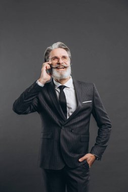 İş ve modaya uygun sakallı, gri takım elbiseli olgun bir adam gri arka planda telefonla gülümsüyor ve konuşuyor..