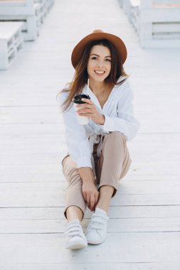 Şık bir kadın kahve içiyor ve gün batımında sıcak bir yaz gününde sahilde yürüyor.