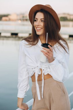 Şık bir kadın kahve içiyor ve gün batımında sıcak bir yaz gününde sahilde yürüyor.