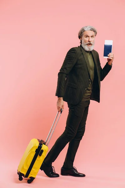 Bearded stylish middle-aged man holding a yellow suitcase and passport with a ticket on a pink background.