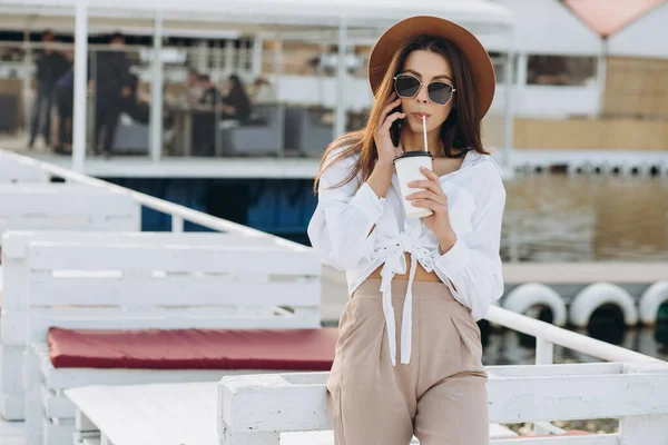 A stylish woman with cup of coffee talking by phone and walk along the beachfront on a warm summer day at sunset