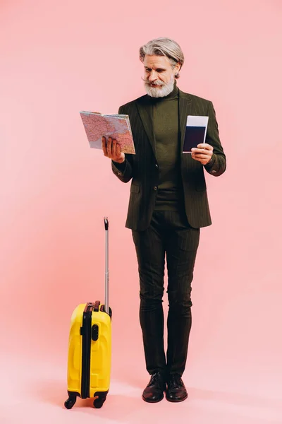 Bearded stylish middle-aged man holding a yellow suitcase, map and passport with a ticket on a pink background.