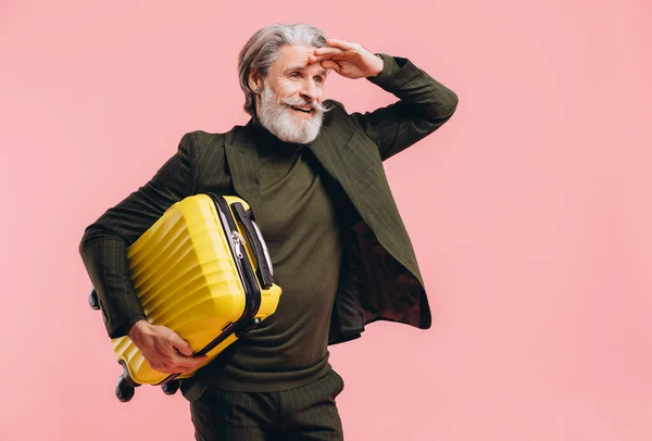 Bearded gray-haired middle-aged man in a suit hold a yellow suitcase on a pink background