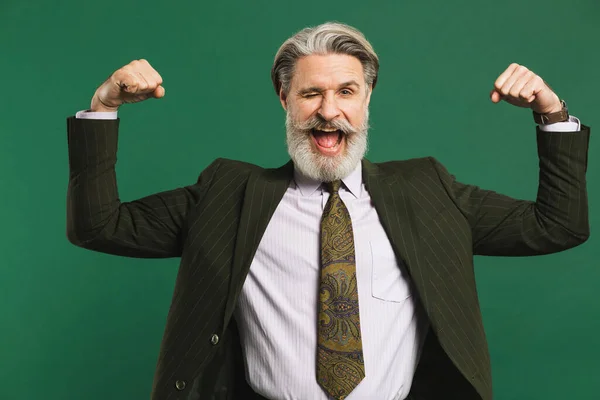 Bearded middle-aged man in khaki suit shows biceps and smiles emotionally on green background