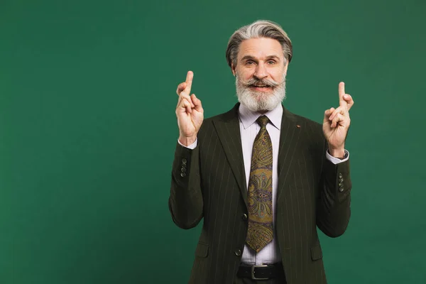 Bearded middle-aged man in suit crosses fingers for good luck on green background.