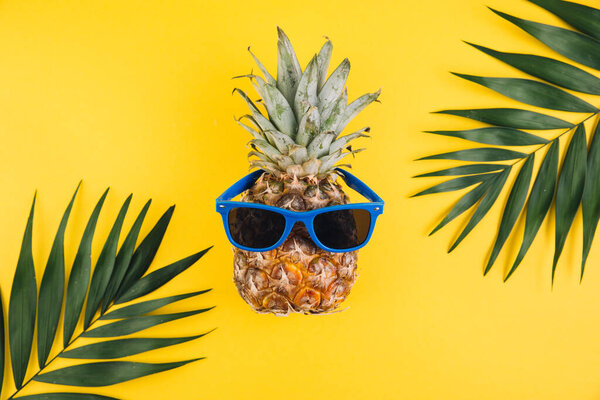 Summer concept. Cute and funny pineapple with sunglasses and palm leaves on yellow background.