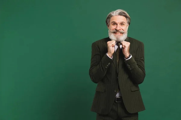 A middle-aged bearded man in a suit holds his fists, aches and rejoices on a green background.