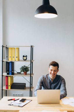 Modern teknoloji ve iletişim konsepti. Geniş dizüstü bilgisayarda çalışan ve ofiste telefon görüşmesi yapan yakışıklı, tıraşsız genç bir iş adamı.