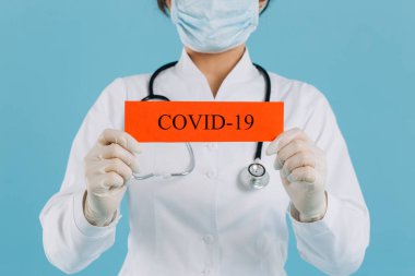 Cerrahi maskeli doktor kadın kırmızı kağıda işaret ediyor. Mesaj Coronavirus, mavi arka planda izole edilmiş.