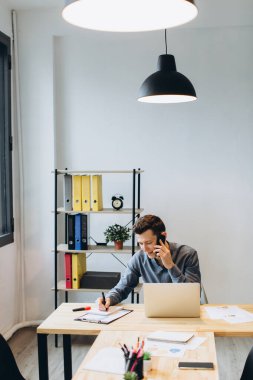 Modern teknoloji ve iletişim konsepti. Geniş dizüstü bilgisayarda çalışan ve ofiste telefon görüşmesi yapan yakışıklı, tıraşsız genç bir iş adamı.