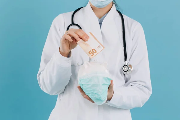 聴診器を装着した医師ピギーバンクとお金は 医療面マスクを着用 コロナウイルスの概念 — ストック写真