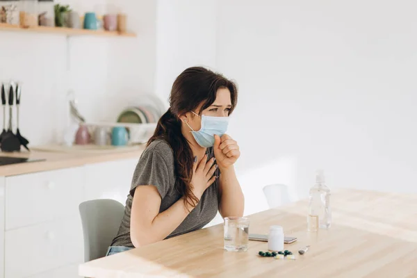 Woman in protective mask coughing in fist sick of coronavirus viral infection spreading coronavirus covering mouth and nose. Painful cough ill patient sitting in kitchen at home quarantine