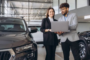 Salesman holding laptop and showing car features to woman in car dealership, explaining automobile specifications