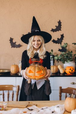 Mother wearing witch costume and daughter holding jack-o'-lantern celebrating Halloween together in decorated home kitchen