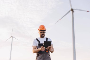 Engineer in a hard hat using a tablet to monitor wind turbines, highlighting sustainable energy solutions and innovation in renewable technology