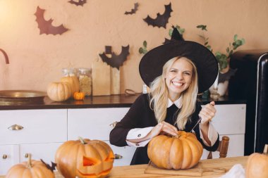 Happy woman wearing witch hat carving pumpkins for Halloween party, enjoying festive preparations in decorated kitchen