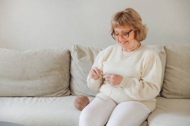 Elderly woman knitting comfortably on the sofa, savoring a peaceful afternoon at home while enjoying her favorite hobby