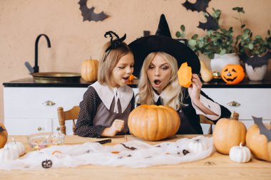 Mother dressed in a witch costume and daughter joyfully carving pumpkins together for a festive Halloween celebration at home