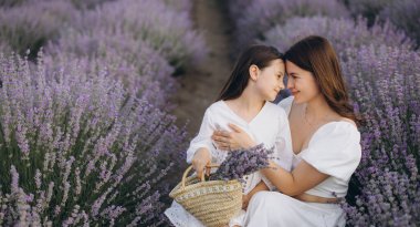 Mother and daughter wearing white dresses are touching foreheads in a lavender field, holding a wicker basket