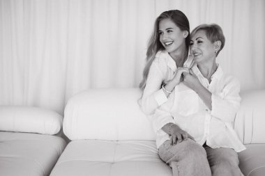 Happy mother and daughter in white shirts smiling and embracing each other while sitting on a white sofa