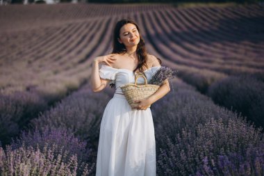 Young woman in white dress holding wicker basket with lavender bouquet in a lavender field at sunset