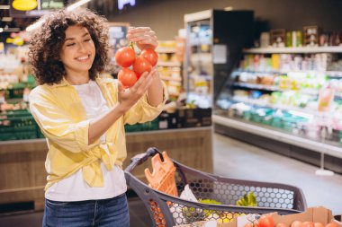 Smiling woman with curly hair selecting ripe tomatoes on the vine while shopping in the grocery store's produce aisle