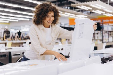Smiling woman opening washing machine checking features in home appliance store, choosing new washer for her apartment