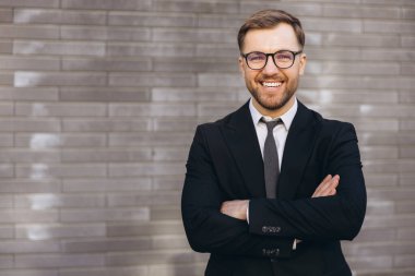 Portrait of a confident businessman wearing glasses and smiling with arms crossed in front of a gray brick wall