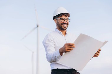 Engineer wearing a hardhat is smiling while reading a blueprint in front of wind turbines, representing sustainable energy and engineering expertise