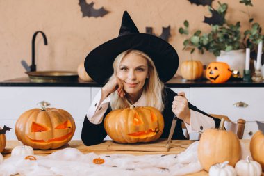 Happy witch carving pumpkins for Halloween party using knife, kitchen decorated with Halloween ornaments