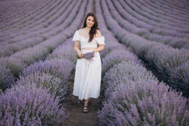 Young woman walking in a lavender field holding a bouquet of lavender flowers, wearing a white top and skirt