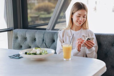 Smiling woman using smartphone at restaurant table with fresh salad and orange juice, enjoying a healthy lunch break