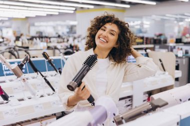 Smiling woman touching her hair and holding a hairdryer in an electronics store, trying different hair styling tools