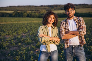 Interracial agronomists couple smiling with crossed arms in cultivated soybean field at sunset