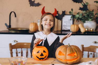 Smiling little girl with devil horns headband and painted face holding a pumpkin bucket celebrating Halloween