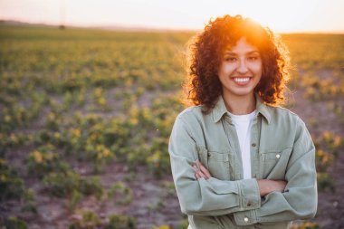 Portrait of a young woman agronomist smiling confidently with crossed arms, enjoying the tranquil beauty of a soybean field at sunset