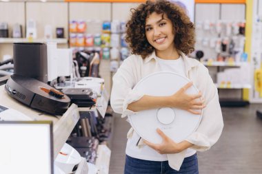 Smiling woman selecting a new robot vacuum cleaner while browsing through the aisles of an electronics store, enjoying the shopping experience