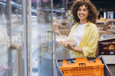 Young woman shopping for fresh eggs in refrigerated aisle of grocery store, holding carton and smiling