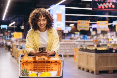 Young woman with curly hair using her smartphone while pushing a shopping cart in a supermarket