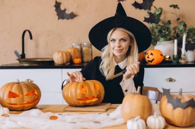 Smiling woman in a witch hat carving pumpkins for a Halloween party, skillfully using a knife in a cozy kitchen filled with festive decor