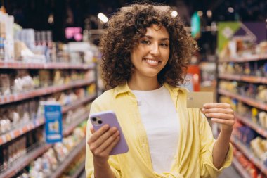 Smiling woman holding smartphone and credit card, enjoying convenient cashless payment while shopping in modern supermarket