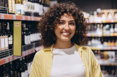 Young curly haired woman smiling while choosing a bottle of wine in a supermarket drinks section