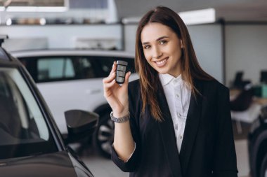 Smiling saleswoman presenting car keys inside a bustling car dealership, showcasing her expertise in customer service and automotive sales
