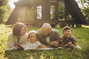 Happy family lying on a blanket in their backyard, enjoying a weekend together in front of their modern a frame house
