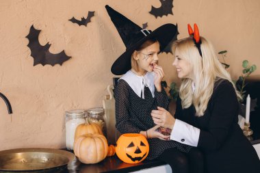 Mother and daughter smiling and having fun wearing Halloween costumes while eating candies in their decorated kitchen