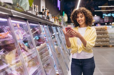 Smiling young woman reading label of packaged meat standing by refrigerated section in grocery store